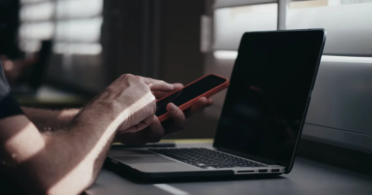 Mains d'une personne tenant un smartphone à côté d'un ordinateur portable ouvert, lumière naturelle filtrée