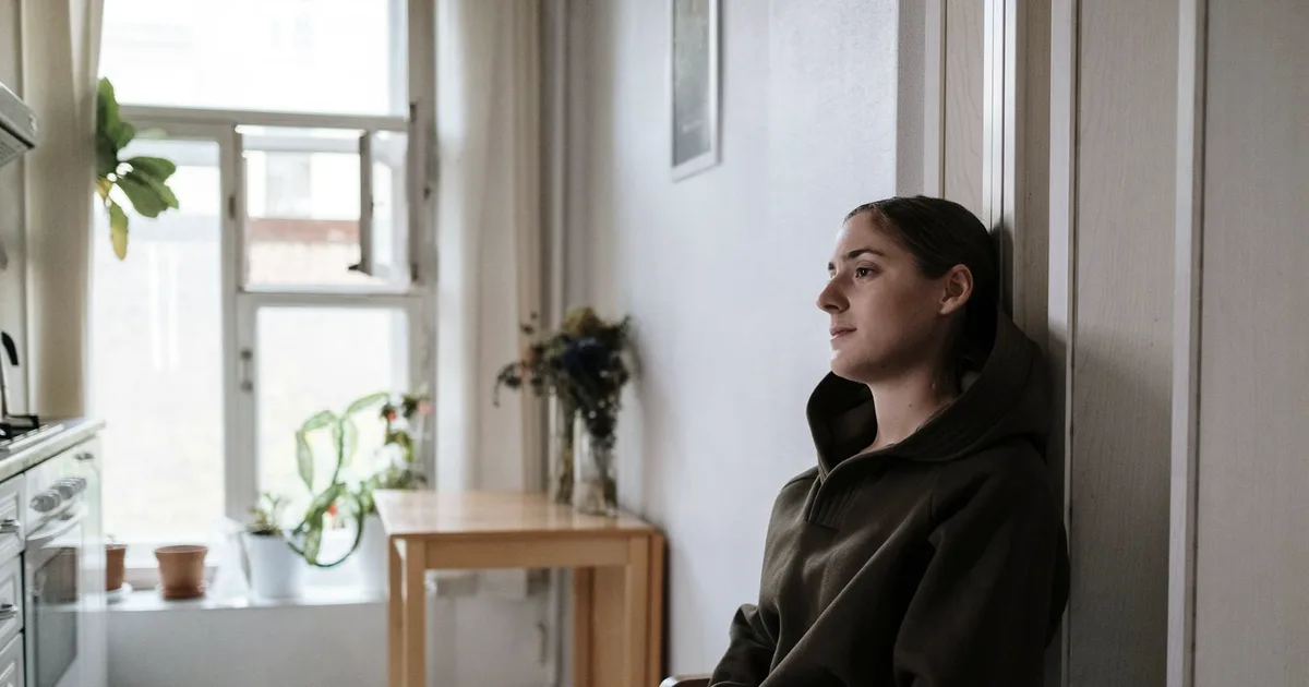 Woman in a hoodie leaning against a wall near a window, gaze elsewhere