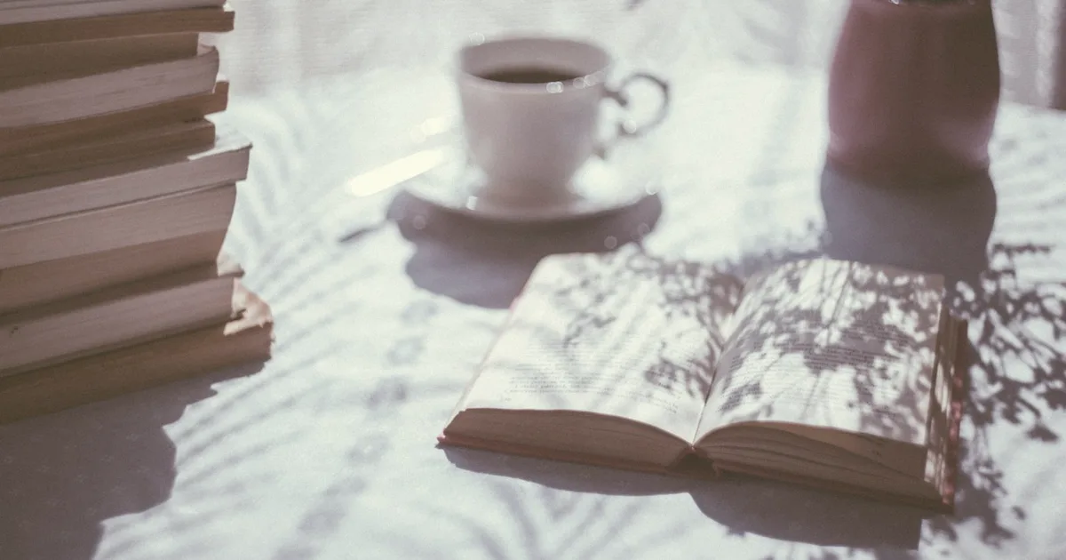 Open book resting on a light-colored table next to a stack of books and a cup, soft natural light