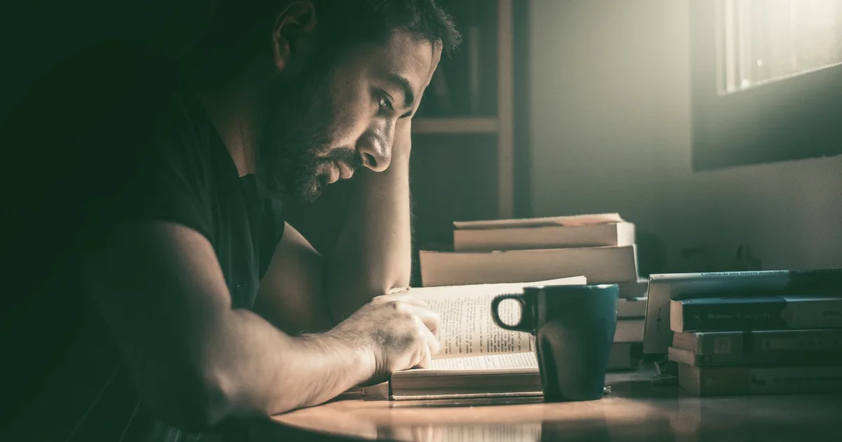 Personne lisant un livre à la lumière chaude d'une lampe, entourée de piles de livres