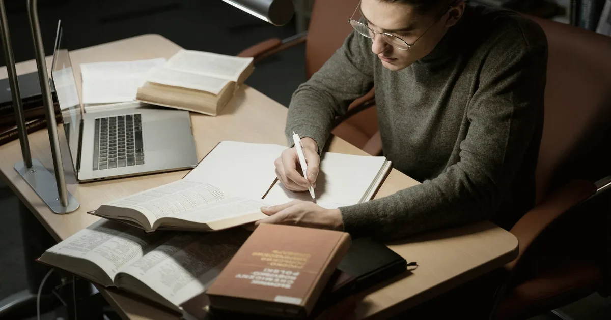 Focused person bent over open books and a notebook under a study lamp