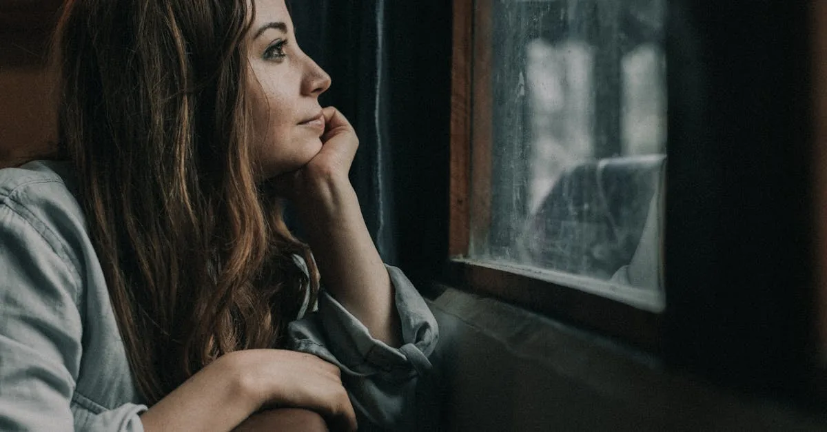 Femme rousse pensive accoudée à une fenêtre, regard tourné vers l'extérieur dans une lumière froide